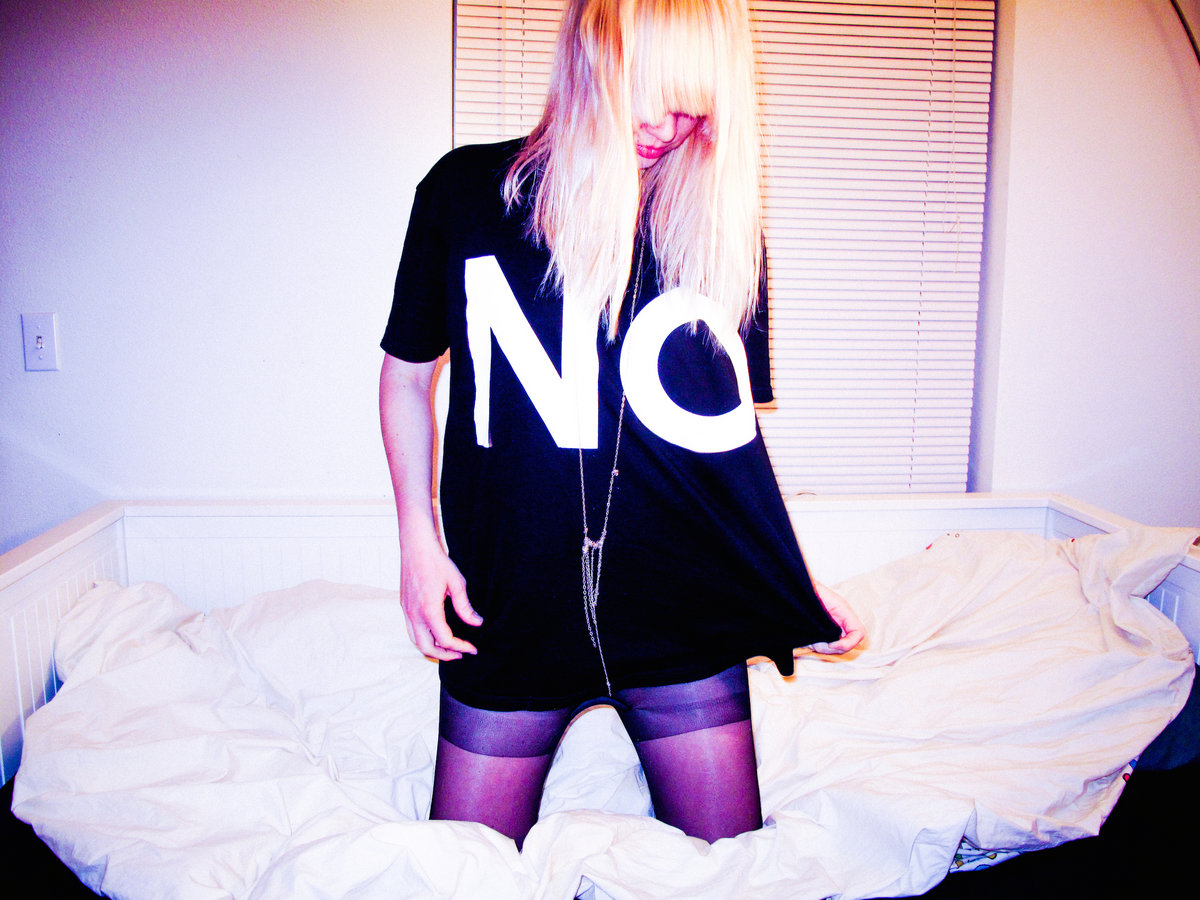 A photograph of EMA kneeling on an unkempt bed with a black t-shirt with big white letters spelling "NO."