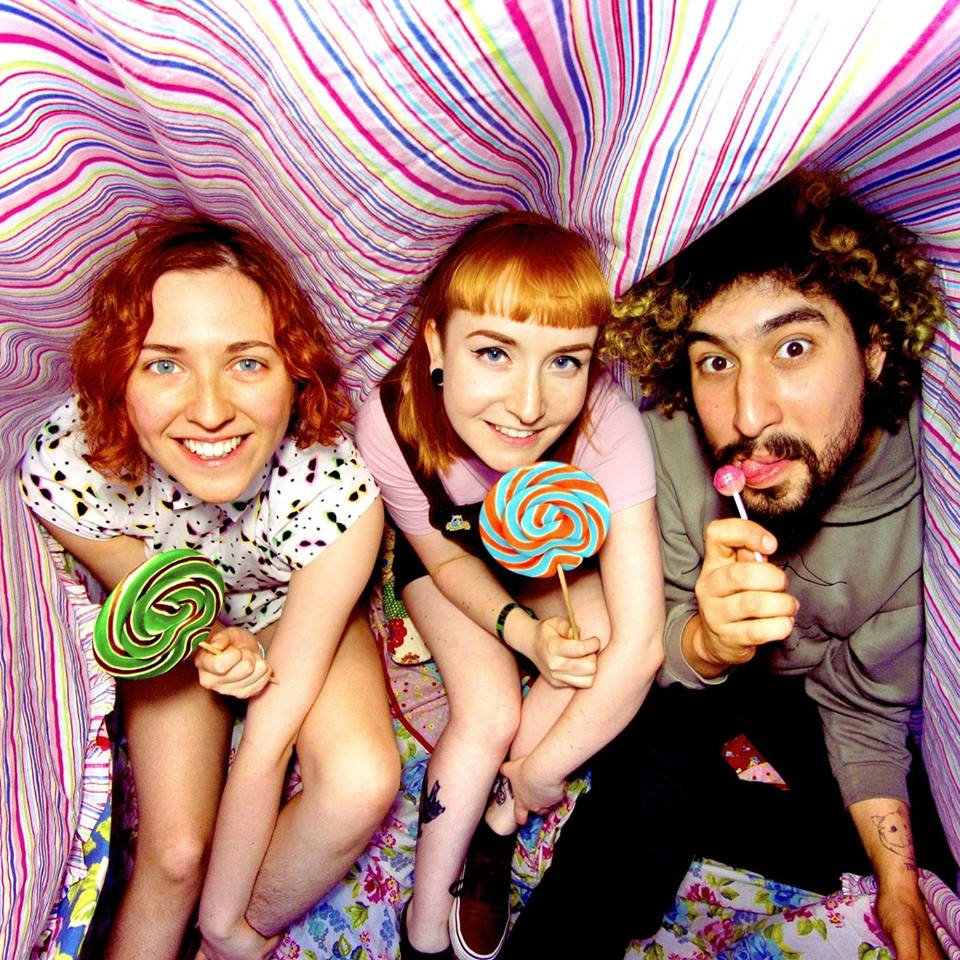Sugar Rush promotional picture. Three people sat in a row under a colourful blanket, each holding a lollipop. The left and middle individuals are smiling and both have red shoulder length hair and blue eyes. They are holding large novelty size lollipops. The individual on the right has slightly shorter than shoulder length curly black hair with bleached ends, and a black beard. They are licking a smaller regular sized lollipop.