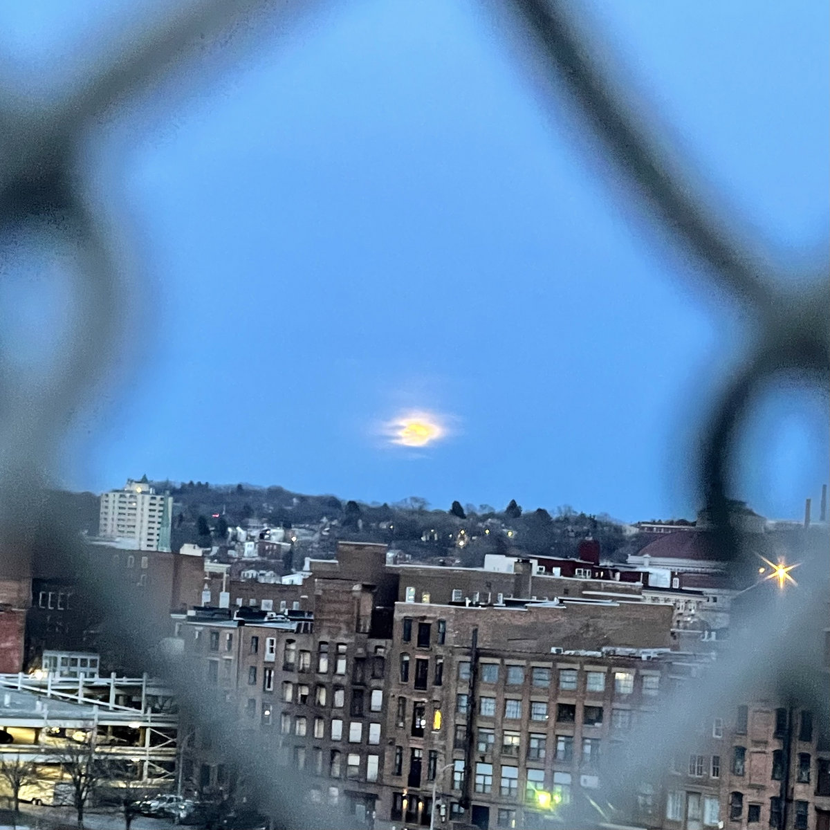 seen through a chainlink fence is a rising moon obscured by clouds over a crumbling victorian town