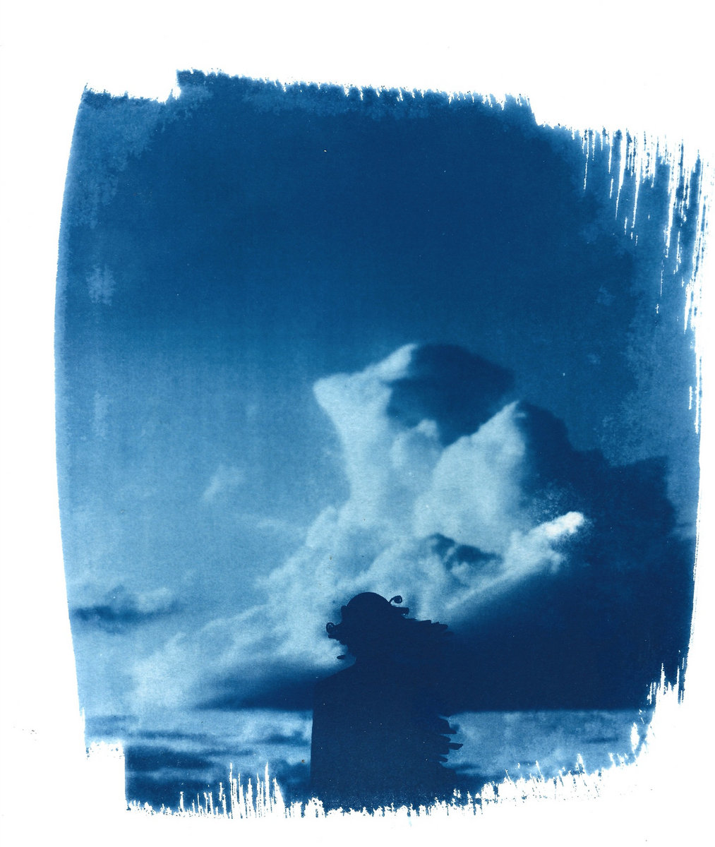 Cyanotype in deep blue with ragged, brushed white edges. A dark silhouetted figure in a crested headpiece stands beneath towering white clouds.
