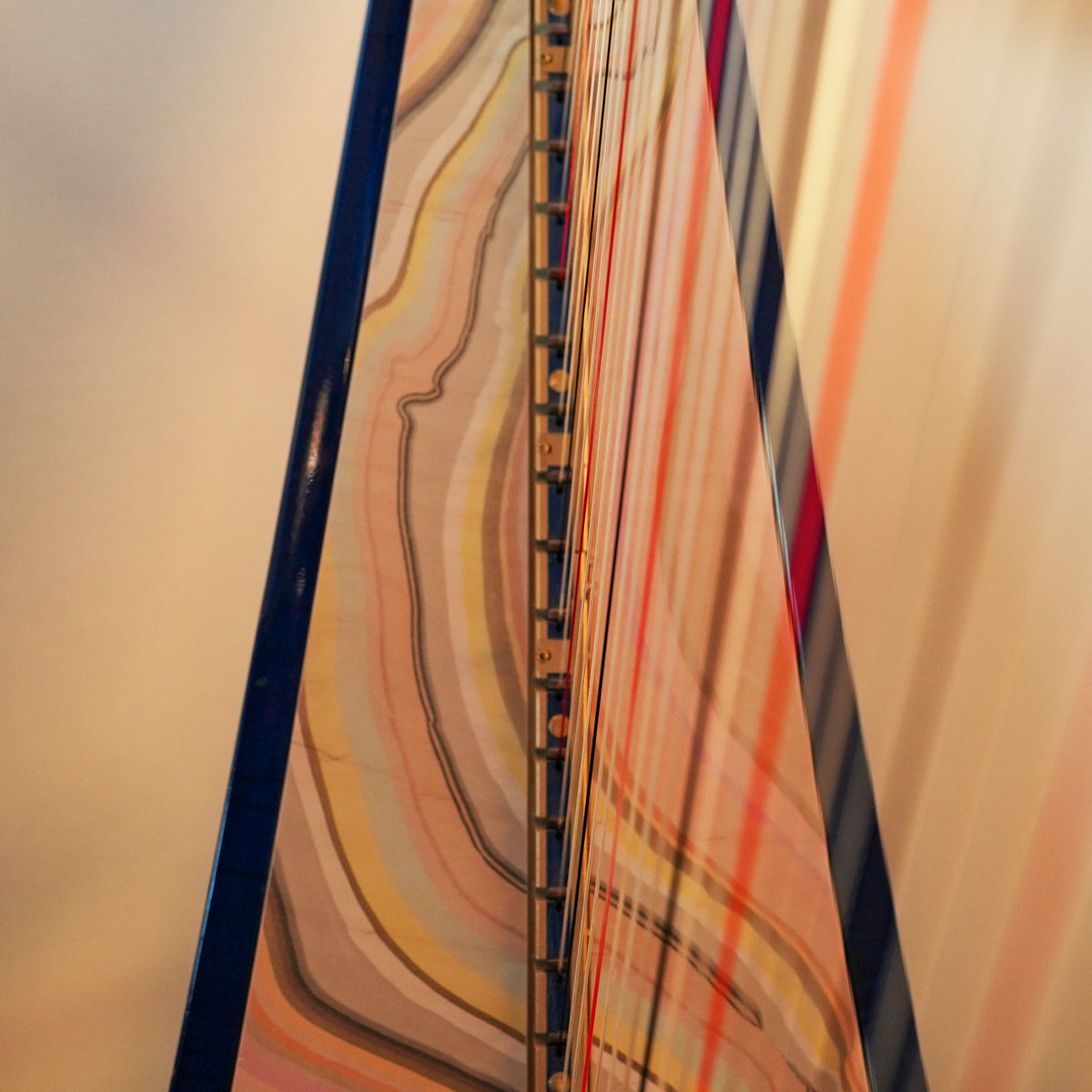 Close-up of a harp rising to a point, its soundboard marbled in bands of peach, yellow, and grey beside a navy column, red and gold strings blurring into a cream background