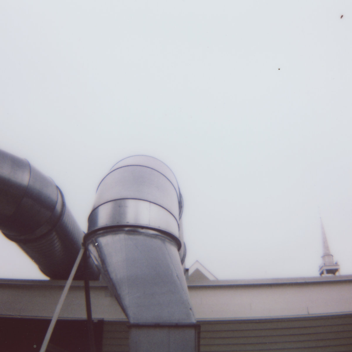 Instant-film photo of galvanized steel ductwork curving up from a rooftop against a pale gray sky, with a white church steeple at the right edge.