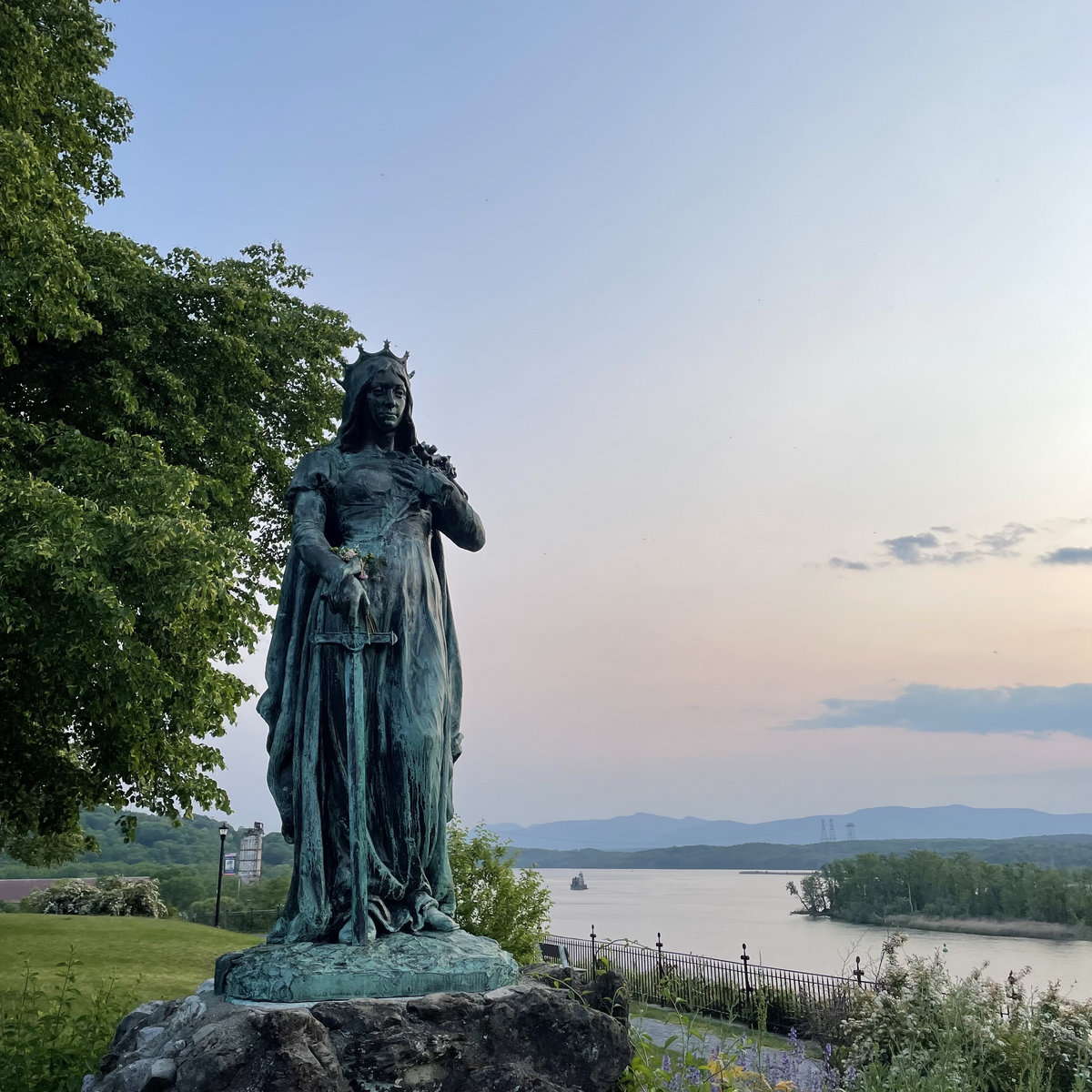 a beautiful copper statue stands majestically above a sunset over the hudson river and there is a tiny little lighthouse in the distance