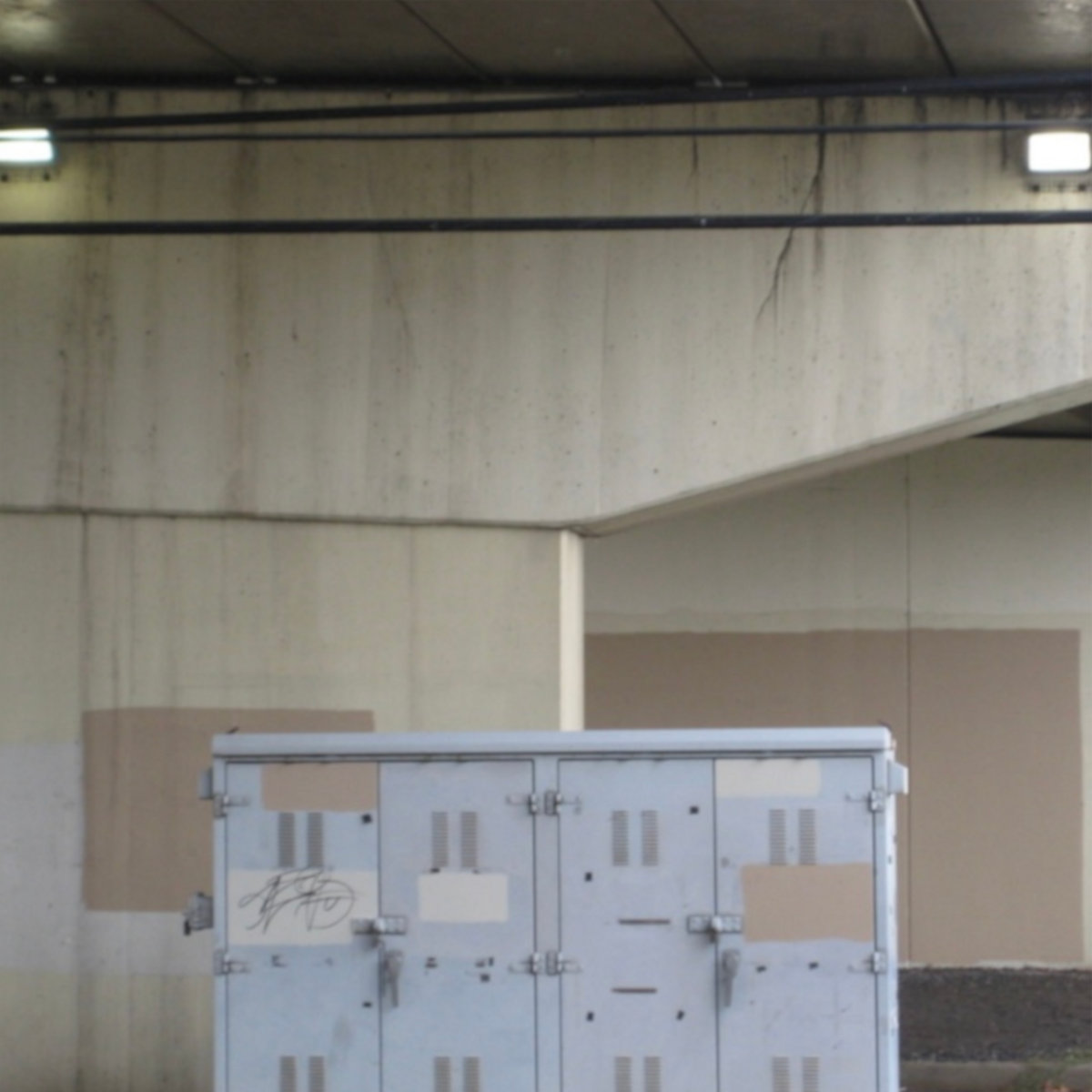 A pale blue-grey metal utility cabinet stands against concrete walls patched with rectangles of beige paint, beneath an overpass ceiling crossed by black cables and two strip lights.